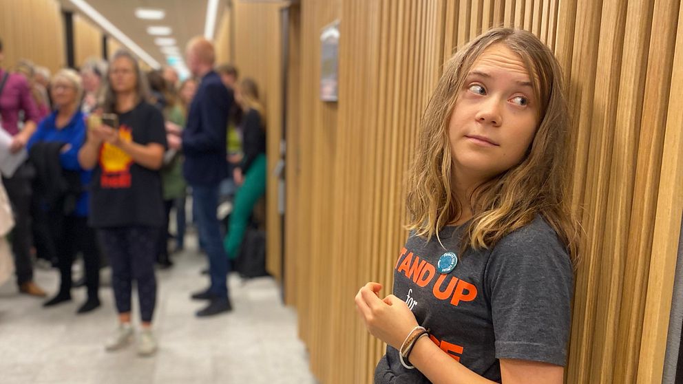 Greta Thunberg lutar sig mot väggen på Malmö tingsrätt till vänster står folk och fotar henne. Greta tittar bort.