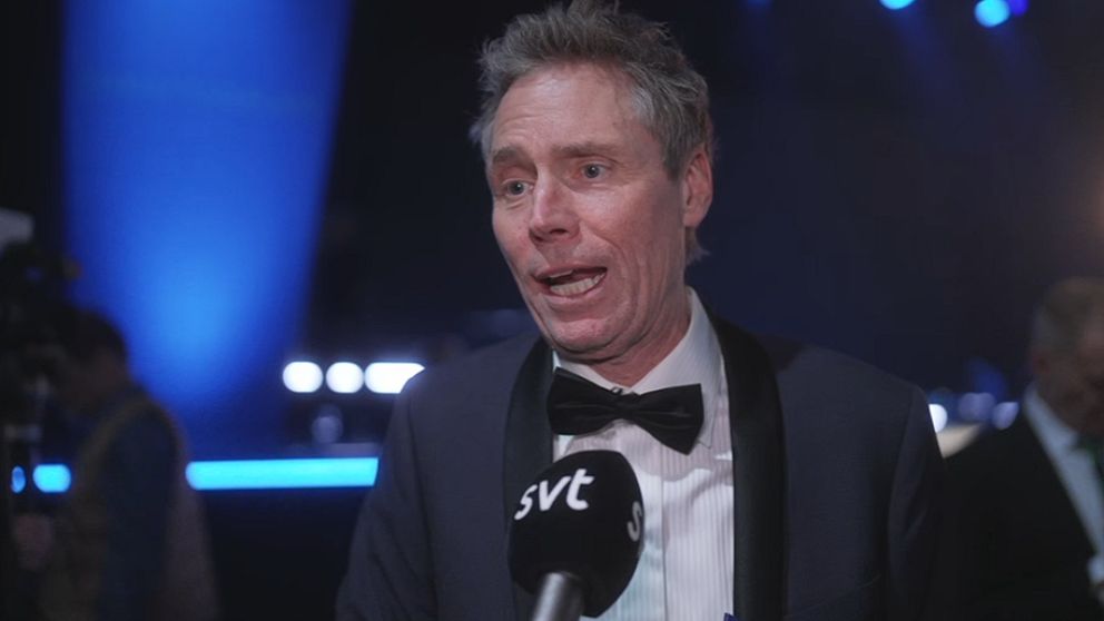 En man i kostym med fluga intervjuas, stående i en scenisk miljö med blå belysning. Bakgrunden visar suddiga konturer av människor och ljus, vilket tyder på ett evenemang eller en gala.
