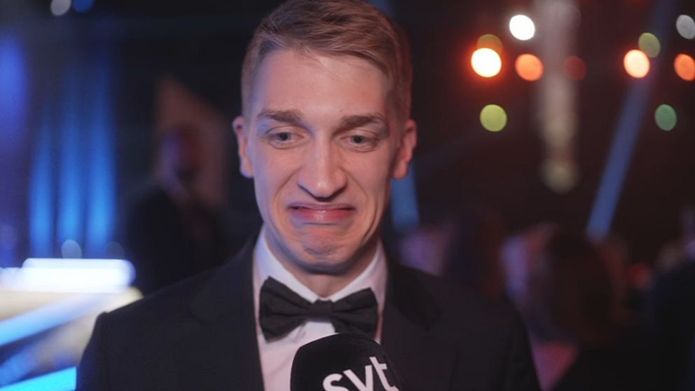 En man i frack ger en intervju med en mikrofon märkt ”SVT” i en festlig miljö med dämpad belysning och oskarpa människor i bakgrunden.