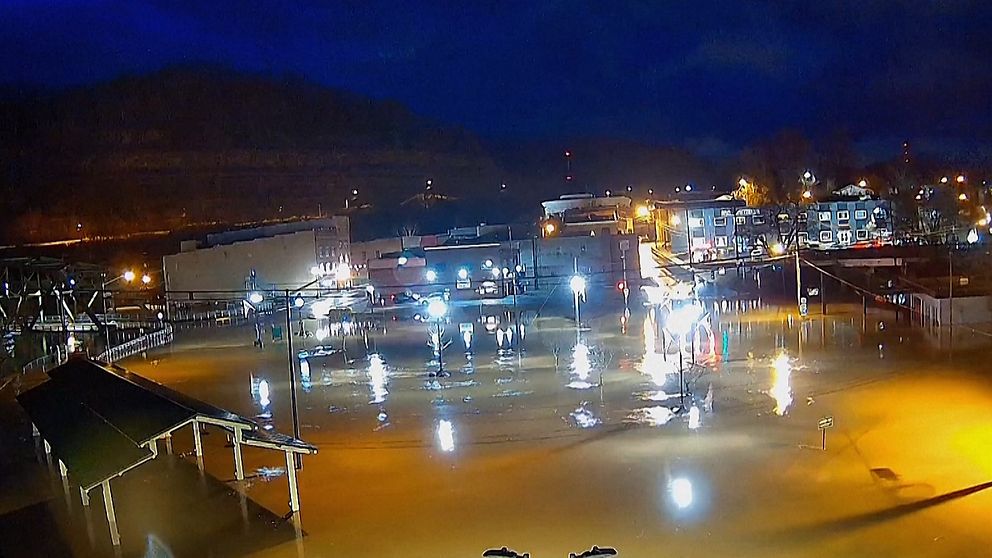 Översvämmat industriområde på natten, med vatten täckande marken och reflekterande ljus från omgivande byggnader och gatubelysning.