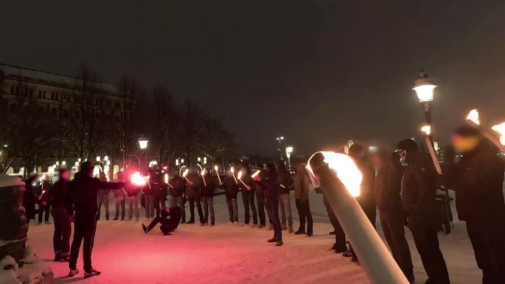 Personer står i ring utomhus på natten och håller facklor, snö på marken.