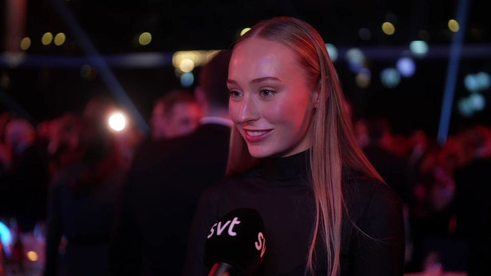 En kvinna i en svart outfit står i fokus för en intervju på en gala. Bakgrunden visar suddiga silhuetter av människor och färgglada ljuspunkter som skapar en festlig atmosfär.