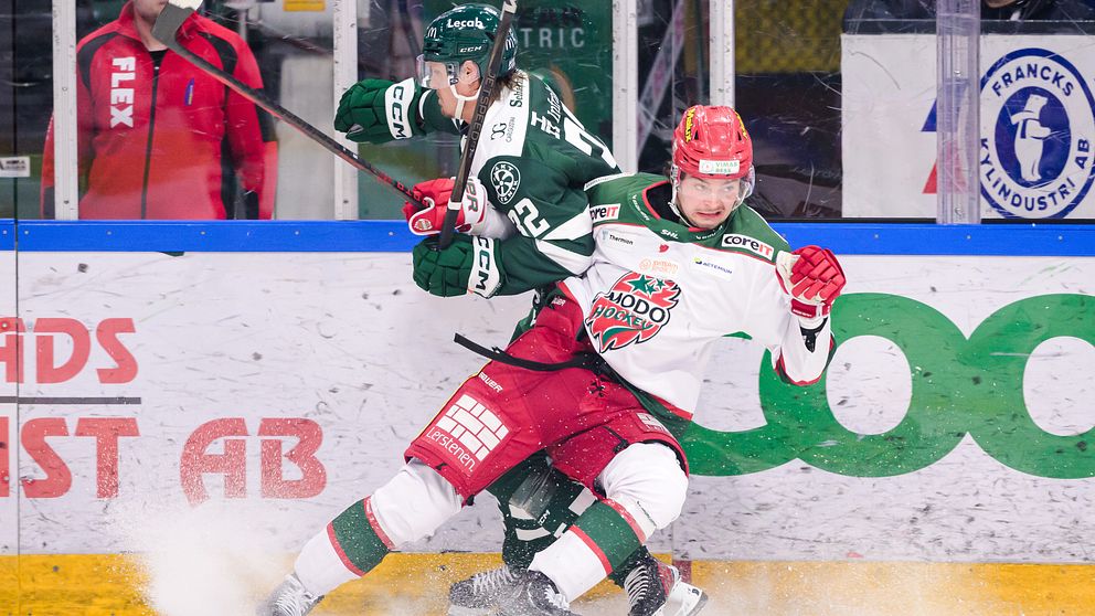 Två ishockeyspelare i intensiv kamp på isen, där en spelare från laget i gröna tröjor blir tacklad av en motståndare i vit och röd tröja. I bakgrunden syns åskådare och delar av ishallarens inredning.