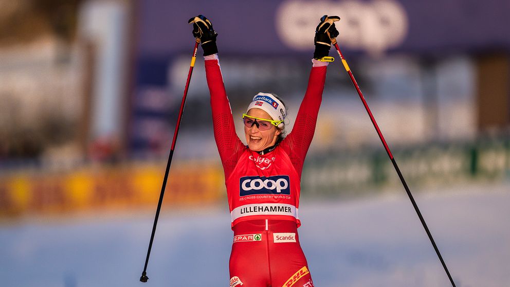 En skidåkare sträcker händerna mot skyn i segergest efter ett lopp i Lillehammer. Personen bär en röd tävlingsdräkt och håller stavarna i händerna. Bakgrunden visar en snötäckt bana och publik.