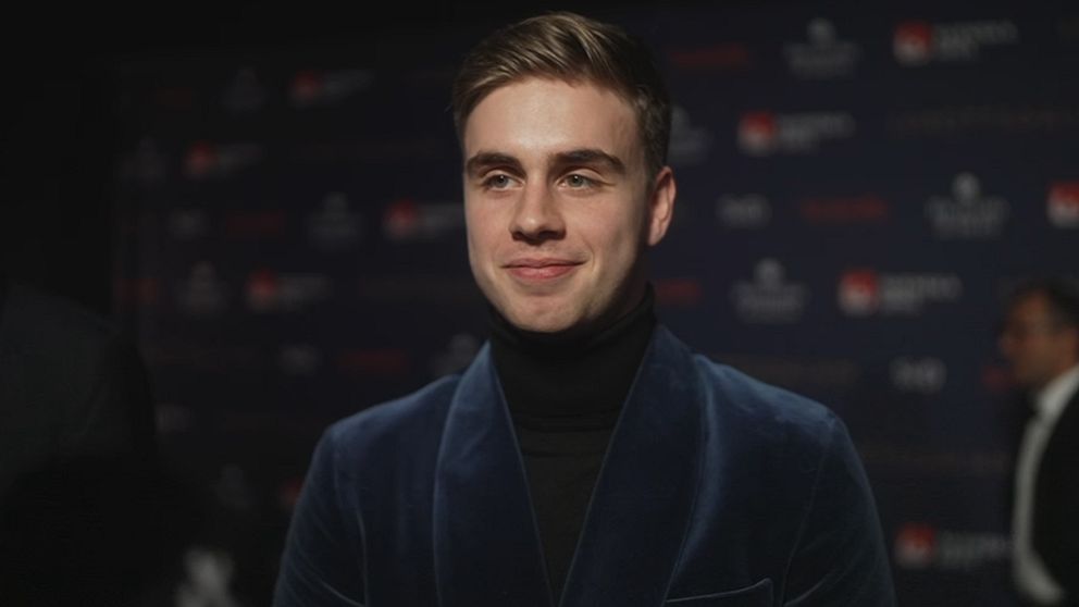 En person iklädd en blå sammet blazer står framför en mörk bakgrund med diskreta logotyper, troligtvis på en gala eller ett offentligt evenemang.