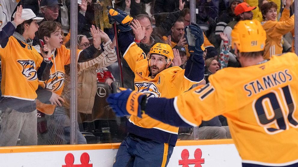 En hockeyspelare i gult lagtröja firar ett mål med utsträckta armar framför en entusiastisk publik som hejar på honom. En annan spelare i gult tröja, med nummer 91, står i förgrunden och vänder sig mot sin lagkamrat för att fira.