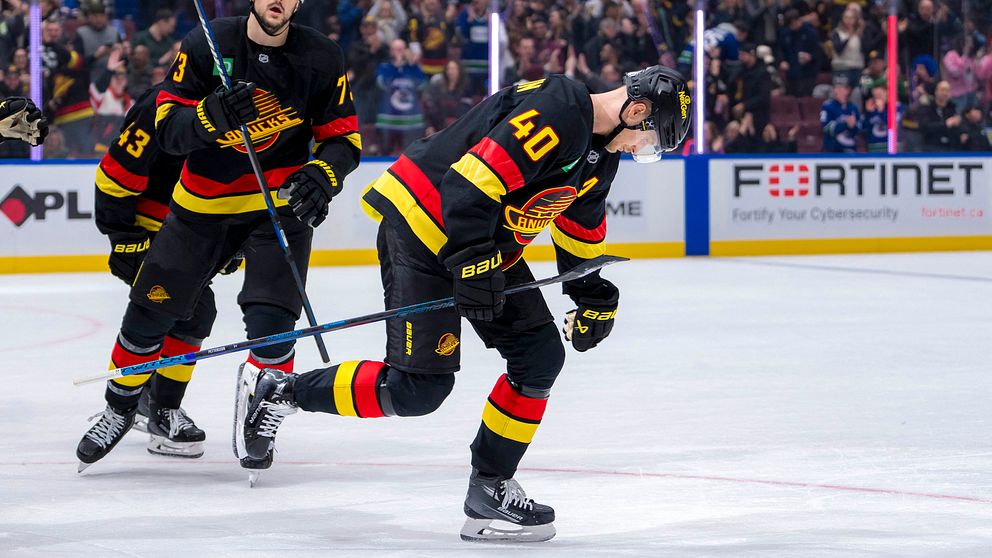 Två spelare från Vancouver Canucks firar efter en målskytt, klädda i lagets färgglada tröjor med röd, gul och svart. Den främre spelaren, med nummer 40, böjer sig framåt med sin klubba upplyft, medan den bakre spelaren, med nummer 73, har en aktiv gest i bakgrunden. Arenan är fylld med åskådare som hejar på i bakgrunden.