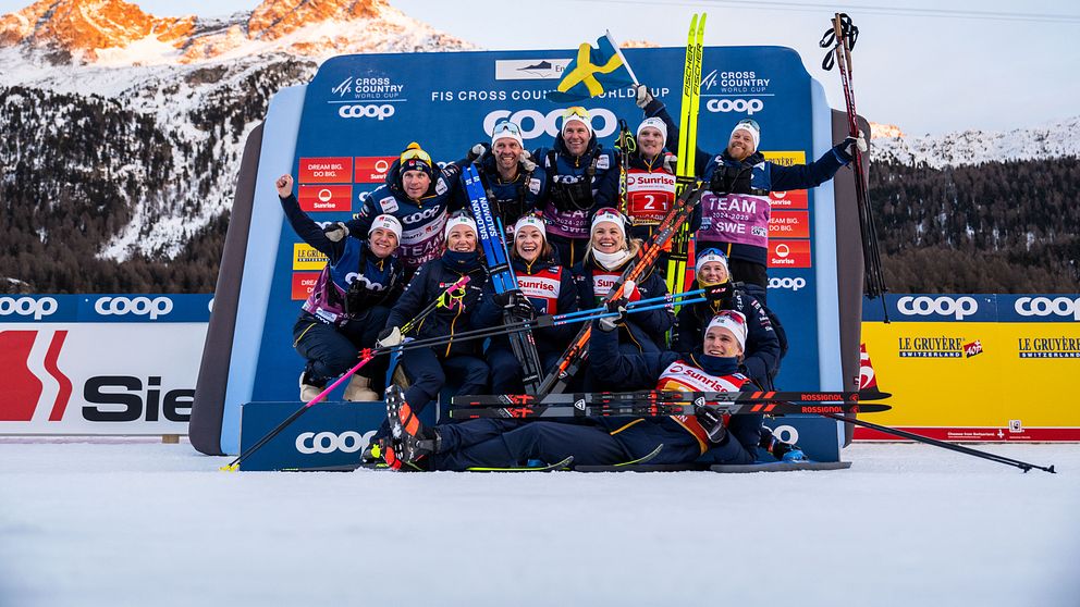 Svenska längdskidlandslaget firar ett lagresultat vid FIS Cross Country World Cup. Gruppen poserar glatt framför ett podium, med skidstavar och en svensk flagga, med snöklädda berg i bakgrunden.