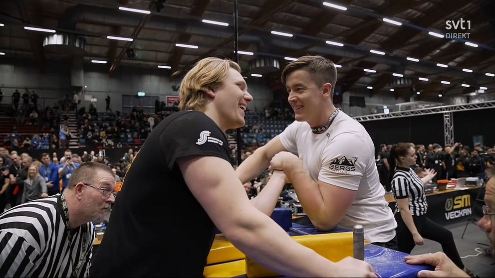 Armbrytningstävling pågår med två deltagare i en arena fylld med åskådare. En domare står nära och håller koll på tävlingen, medan det står ”SM VEDERV” på sidan av bordet, vilket antyder att det kan vara en del av en större tävling eller mästerskap.