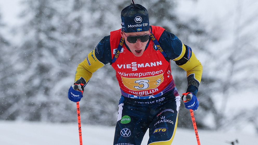 En skidåkare i färgglada tävlingskläder tar sig fram på snön med stavar i händerna, medan åskådare syns i bakgrunden under en vintersporttävling.