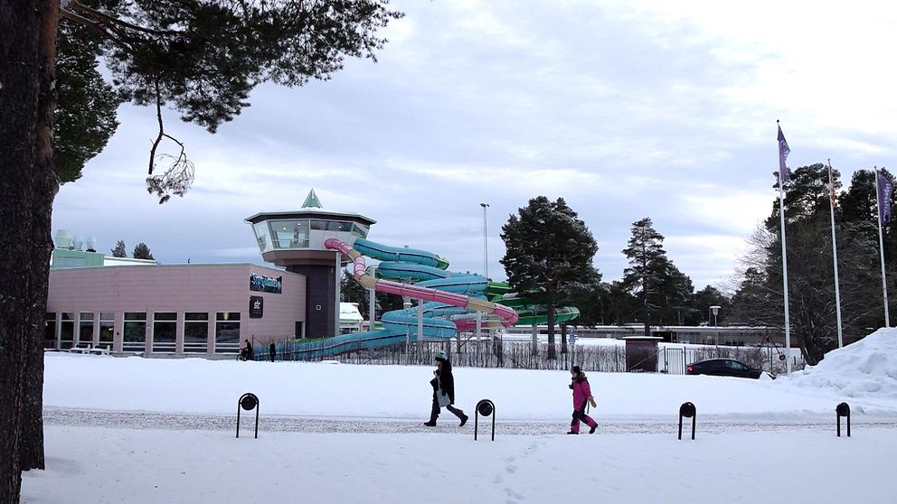 En vintrig scen med snötäckt mark där två personer går på en väg. I bakgrunden syns en vattenpark med färgglada rutschkanor och en byggnad i ljust rosa. Träd och flaggor är synliga på platsen, vilket ger en känsla av friluftsliv.