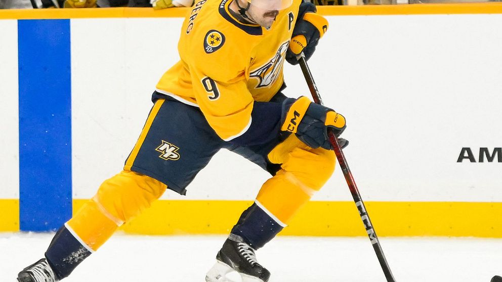 En ishockeyspelare i en gul tröja utför ett skott med pucken under en match, medan lagkamrater och motståndarlaget syns i bakgrunden.