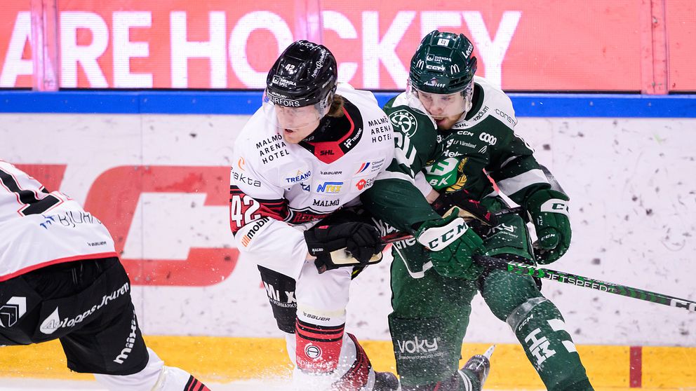 Två ishockeyspelare är engagerade i en närkamp på isen medan en tredje spelare observerar från vänster. Den ena spelaren bär en vit tröja med röd och svart detalj, medan den andra har en grön tröja. Bakgrunden visar en stor reklamskylt som annonserar för ishockey.