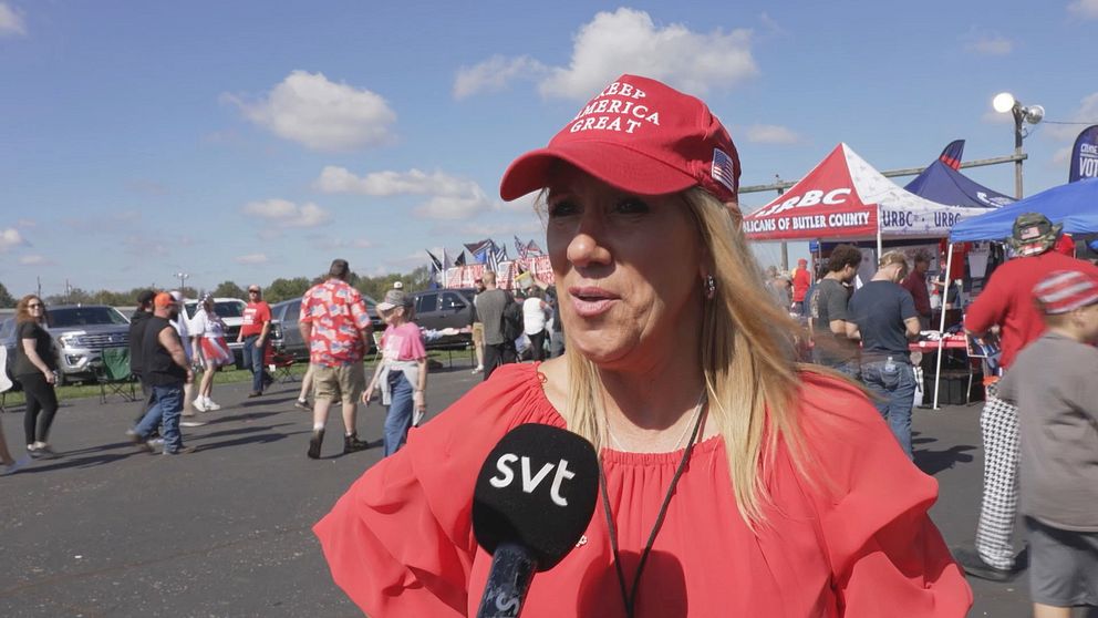 En person i röd keps med texten ”Make America Great Again” står framför en folkmassa vid en utomhusövning. Bakgrunden visar tält, flaggor och besökare, som samlas i en festlig atmosfär under en klar himmel. Mikrofonen är riktad mot personen som verkar delta i en intervju.
