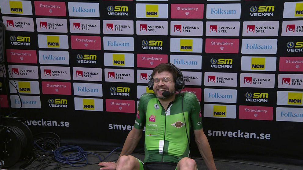 En cyklist i grön tävlingsdräkt sitter framför en sponsorbakgrund med logotyper från olika företag som Folksam, SM-Veckan och Svenska Spel, samt sportrelaterade bolag. Runt cyklisten ligger elektriska kablar.