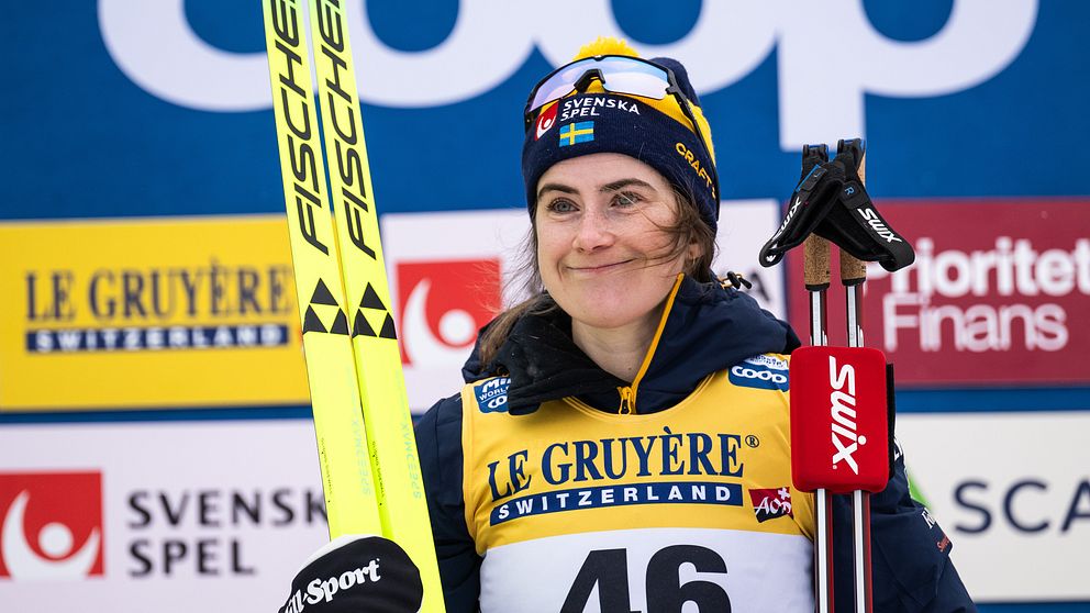 En skidåkare klädd i tävlingsdräkt med nummer 46 och texten ”FALUN” synlig på bröstet står med sina skidor och stavar. Bakgrunden består av olika sponsormärken.