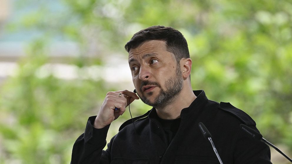 Ukrainas president Volodomyr Zelenskyj talarstol utomhus lyfter handen mot hakan, grönskande bakgrund.