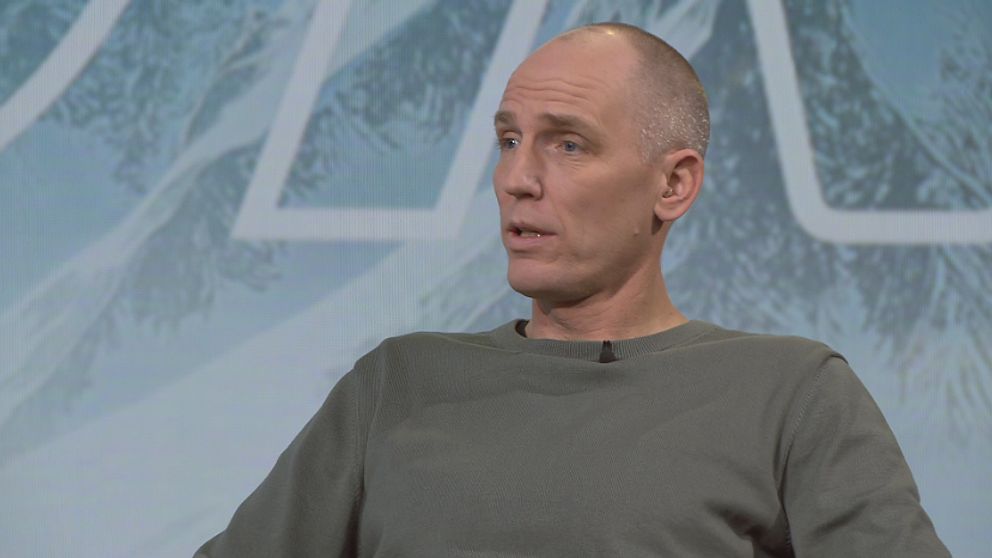 En person sitter på en scen iförd en grå tröja. Bakgrunden visar en stiliserad vinterlandskap med snöklädda berg. Mötet verkar vara en del av en presentation eller diskussion.