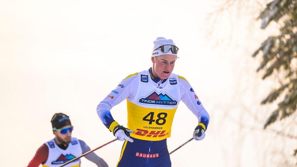 Två skidåkare tävlar i en snöig miljö. Den främre skiljer sig genom att bära nummer 48 på sin tävlingströja, medan den bakre har nummer 43. Båda är klädda i typiska tävlingskläder och använder stavar för att navigera spåren. Det ser ut att vara en solig dag, med snö som glittrar i bakgrunden.