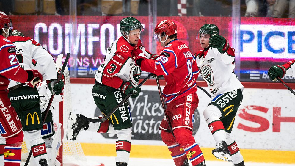 Två ishockeylag är i en intens kamp på isen. Spelare i röda och gröna uniformer är i fokus, med en spelare som firar ett mål och en annan som sträcker sig fram för att gratulera. På andra sidan syns spelare i vita uniformer som försöker försvara sig. Atmosfären är laddad och åskådarna i bakgrunden bidrar till den spännande stämningen.
