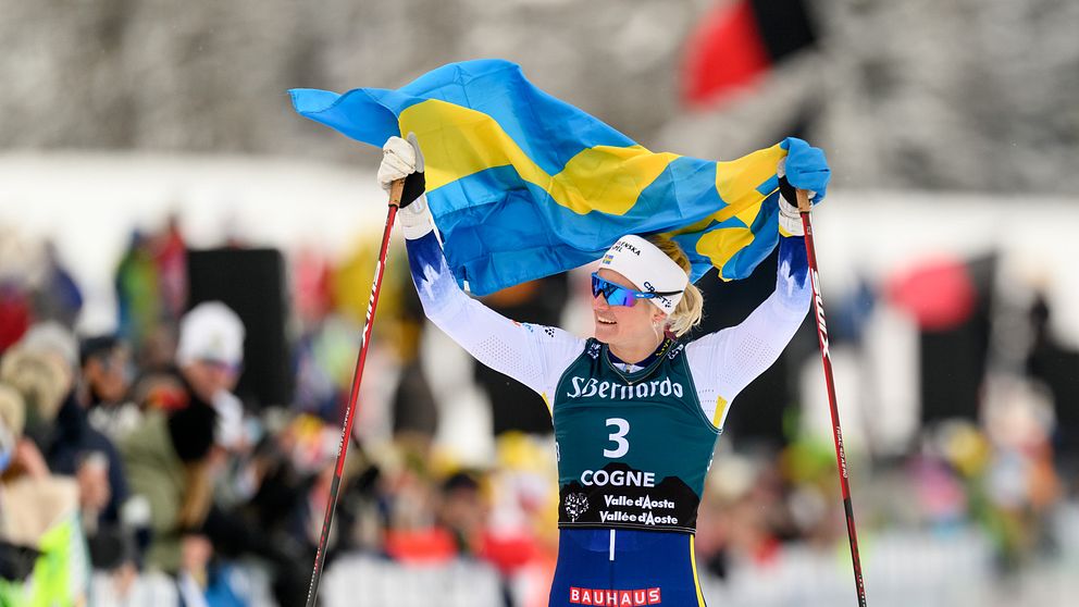 En längdskidåkare iklädd nummerlapp 3 från Cogne, håller en svensk flagga ovanför sitt huvud efter ett tävlingslopp, med en publik i bakgrunden.