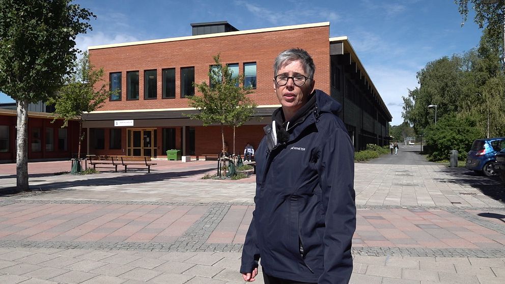 Kvinna med grått kort hår och glasögon står framför Lugnviksskolan, en röd tegelbyggnad.