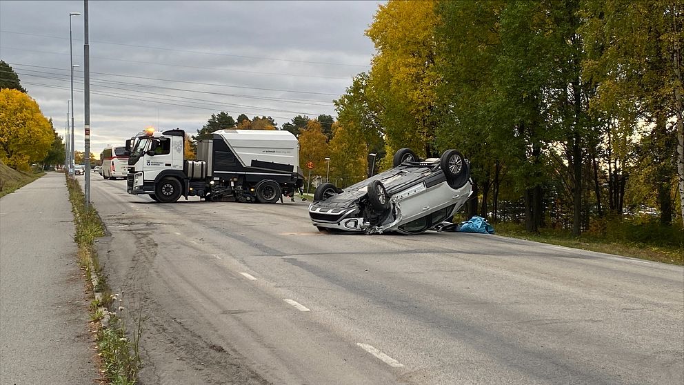 En bil ligger upp och ner, med en sopbil i bakgrunden. Omgivningen är präglad av höstfärger med träd i gult och grönt. Vägbanan är delvis blockerad av den olyckade bilen.