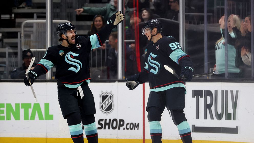 Två spelare från Seattle Kraken firar ett mål på isen, iförda svarta tröjor med turkosa detaljer. Den ena spelaren sträcker upp sina armar i triumf medan den andra ler emot honom. I bakgrunden syns en entusiastisk publik som hejar på.