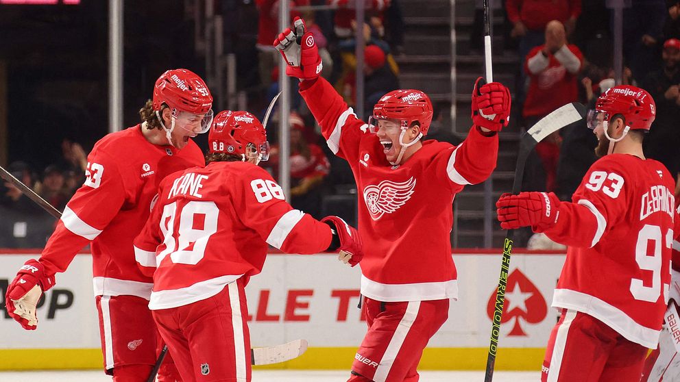 Fyra spelare från Detroit Red Wings firar en seger på isen, klädda i sina röda tröjor. En spelare lyfter armarna i triumf medan de andra ler och klappar om honom. I bakgrunden syns en stor skylt som meddelar ”RED WINGS WIN”.