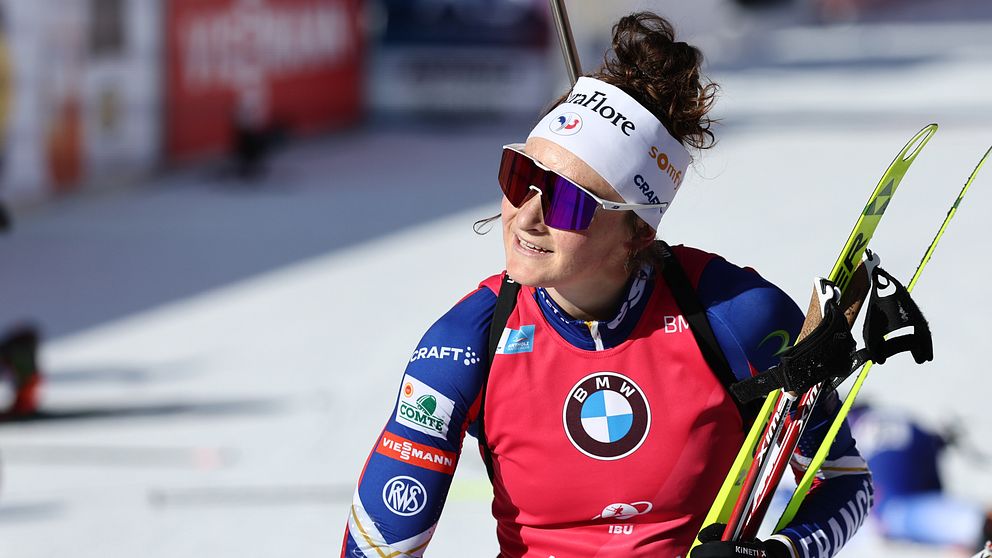 En kvinnlig skidskytt i tävlingsdräkt ses på skidspåret bärandes en gevär och stavar. Hon har en röd nummerlapp med sponsormärken och en vit pannband.