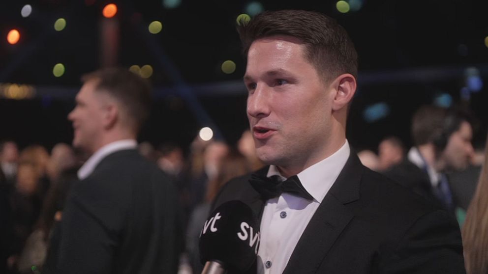 En man i kostym står framför kameran och intervjuas av en reporter med en mikrofon märkt ”SVT”. Bakgrunden visar en kvällsevenemang med suddiga silhuetter av andra gäster och färgglad belysning.