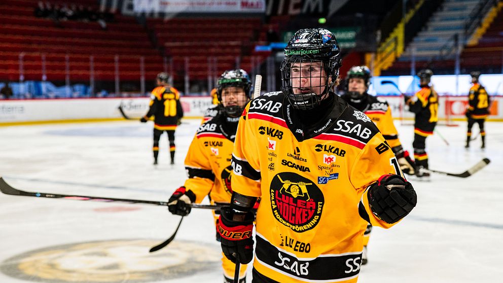 En grupp hockeyspelare i gula tröjor med lagets logotyp och sponsormärken, som rör sig på isen i en ishockeyarena. Bakgrunden visar tomma läktare, vilket tyder på en pågående träning eller match för laget. Spelarna verkar fokuserade och engagerade i aktiviteten.