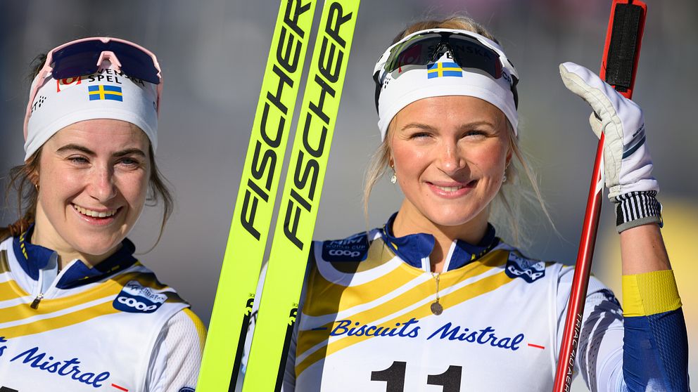 Två skidåkare i svenska landslagsdräkter håller i sina skidor och stavar, med ”Fischer” synligt på skidorna. De har båda nummerlappar och står på en skidtävlingsplats.