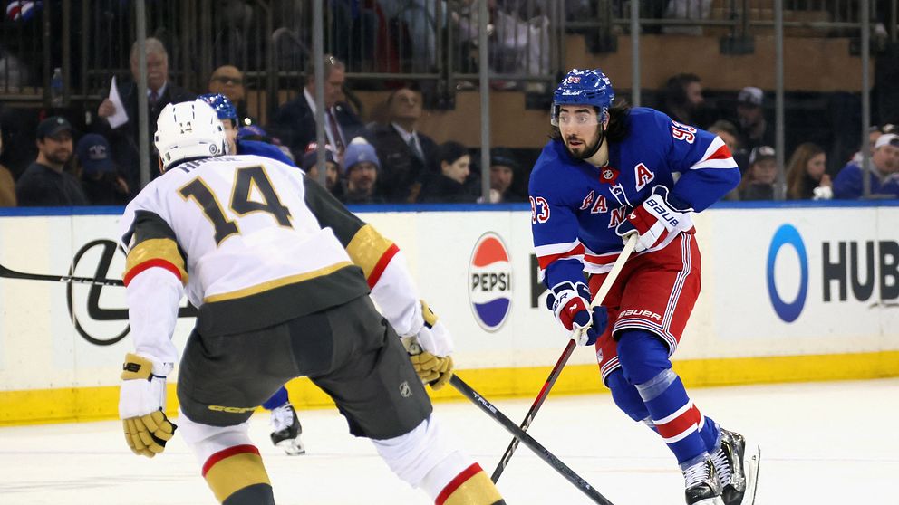 Två ishockeyspelare befinner sig på isen under en match; en spelare i New York Rangers blå uniform manövrerar pucken medan en spelare från det motstående laget i en grå och svart uniform konvergerar mot honom. I bakgrunden syns reklam för Pepsi och Hub.
