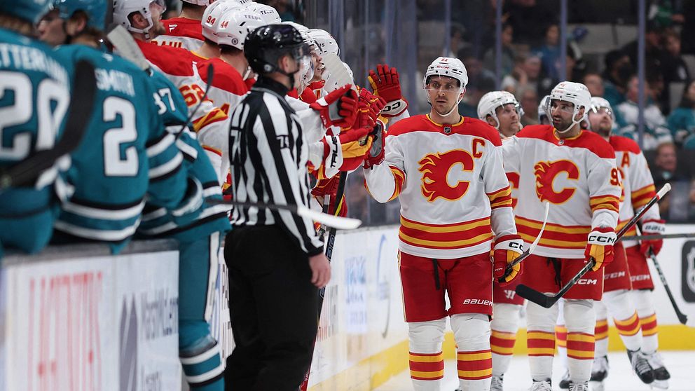 En hockeyspelare från Calgary Flames firar med sina lagkamrater vid bänken efter ett mål under en match. I förgrunden syns en domare i svart-vitt, medan spelare i blått från motståndarlaget San Jose Sharks står på sin bänk i bakgrunden.
