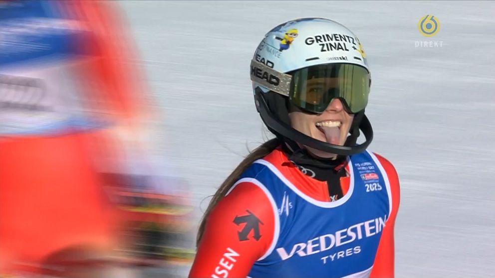 En alpin skidåkare klädd i röd tävlingsdräkt och vit hjälm ler brett med tungan ute under ett lopp. Skidåkaren bär glasögon och i bakgrunden syns en suddig bild av en annan åkare.