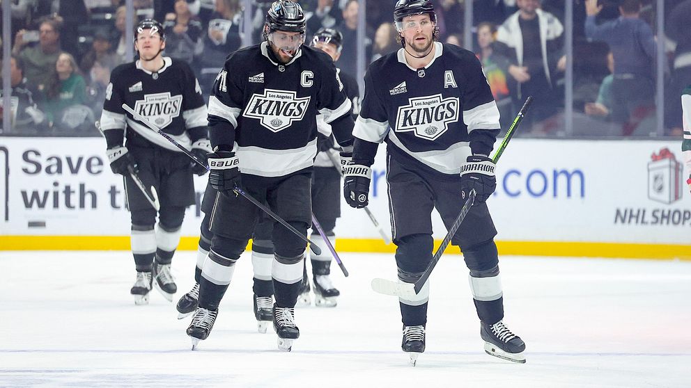 En grupp ishockeyspelare i svart och vitt laguniform går på isen under en match, med publiken i bakgrunden. Spelarna verkar fokuserade och rör sig mot bänken efter en spelsekvens.