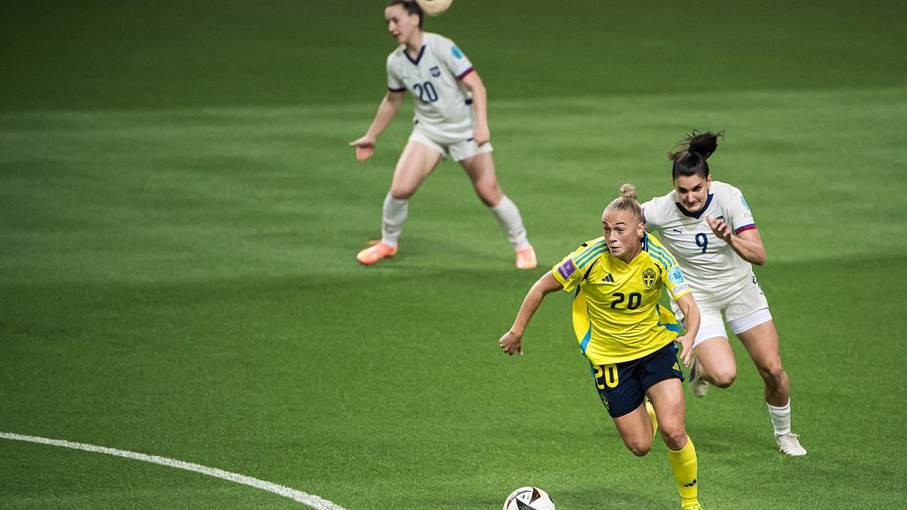 En spelare i gult dribblar bollen framåt på planen under en fotbollsmatch, medan en motståndare i vitt försöker nå henne bakifrån. Gräset är grönt och det ser ut att vara en intensiv situation i spelet.