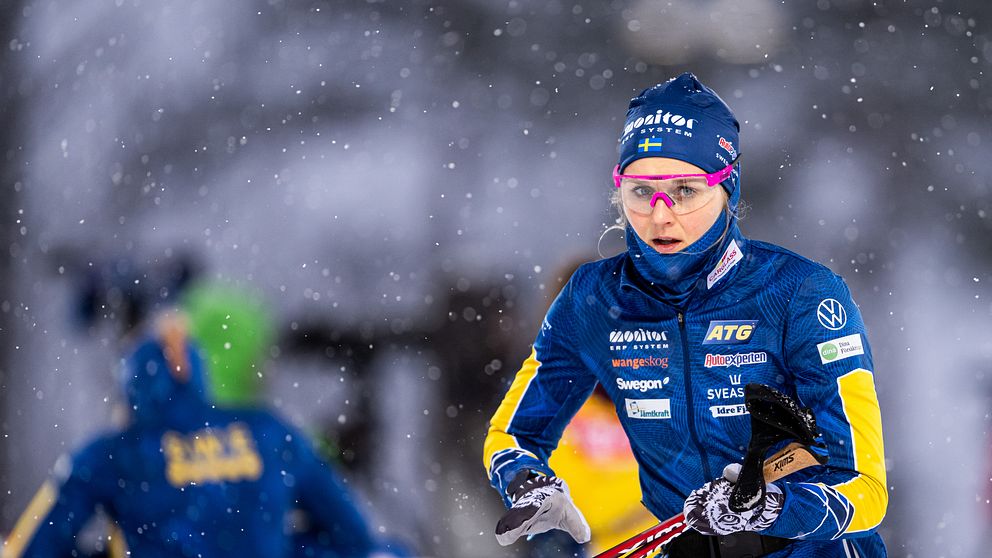 En skidskytt i blå tävlingskläder övar i snöigt väder. Personen har stavar i händerna och är fokuserad på sin teknik. I bakgrunden syns andra personer i tävlingskläder som förbereder sig, vilket förstärker den tävlingsinriktade atmosfären.