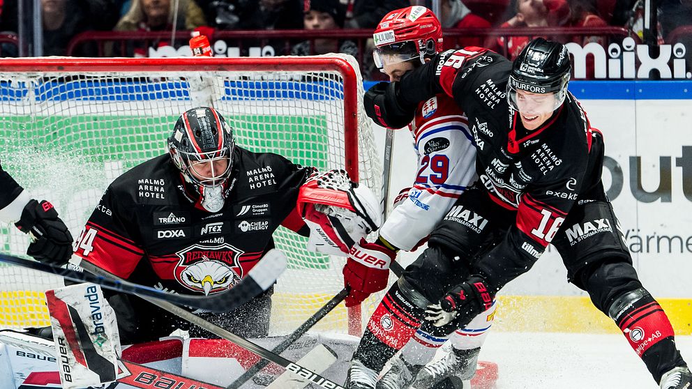 Ishockeyspelare från två olika lag kämpar om pucken nära målet. Målvakten i svarta kläder försöker avvärja ett skott, medan en medspelare skyndar till undsättning. En spelare från det andra laget i vit tröja är också involverad i situationen.