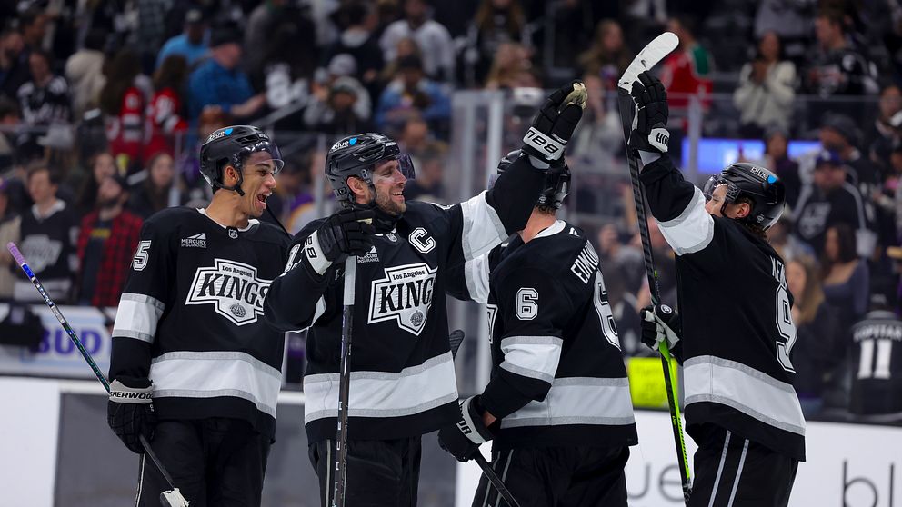 Fyra spelare från Los Angeles Kings firar ett mål på isen, iförda svarta tröjor med lagets emblem. Publiken i bakgrunden är entusiastisk och klädd i olika lagfärger.