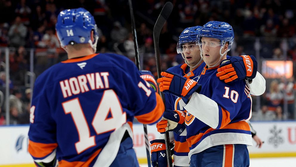 Spelare i orange och mörkblå tröjor firar ett mål på isen under en hockeymatch. Den första spelaren har nummer 14 på ryggen och namnet ”HORVAT”. De andra spelarna omfamnar en lagkamrat, vilket visar en känsla av gemenskap och glädje efter en framgångsrik spelaktion. Bakgrunden visar en publik i arenan.