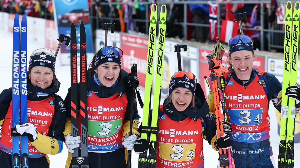 Fyra längdskidåkare i teamkläder poserar med sina skidor och stavar efter en tävling, omgivna av en livlig publik och sponsorfyllda bakgrunder.