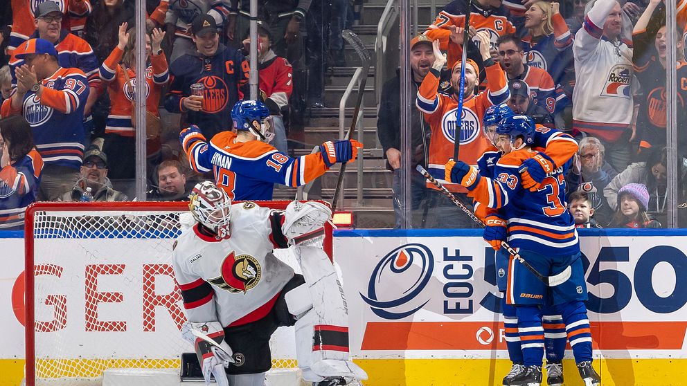 Ett hockeylag firar ett mål på isen medan målvakten från motståndarlaget, som sitter på knä, ser besviken ut. I bakgrunden syns en entusiastisk publik som hejar på laget.
