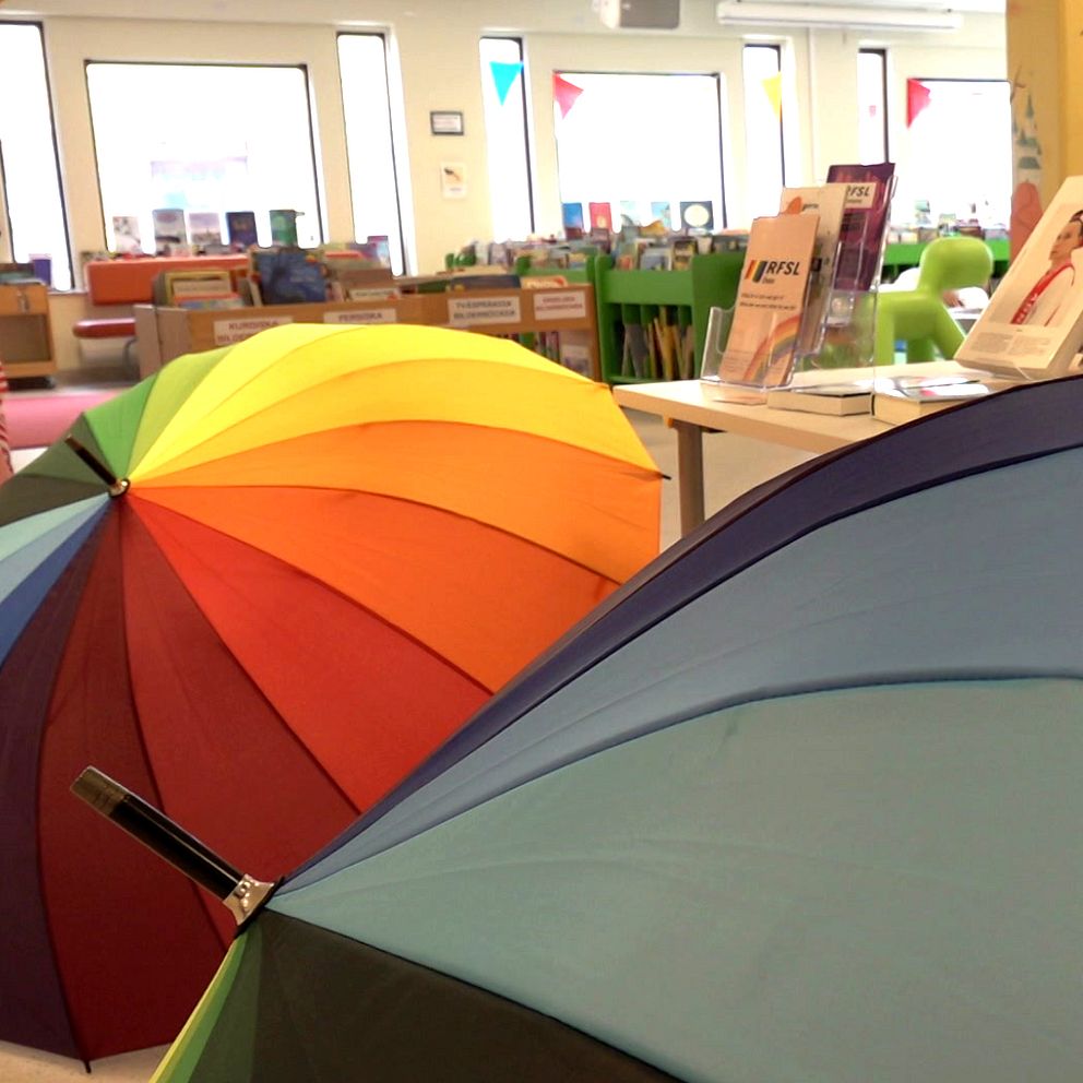 Bibliotek i Lindesberg, två regnbågsfärgade paraplyer i förgrunden samt bild på en man med rakat hår och svarta glasögon.