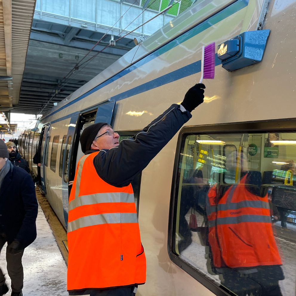 Man i reflexvästar borstar bort snö från kamera på pendeltåg.