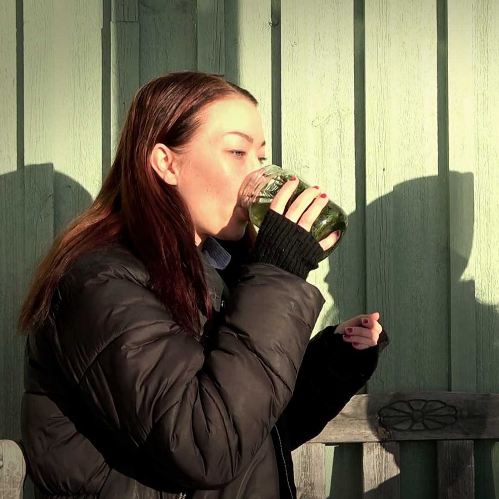 Två personer sitter på en bänk utomhus, en av dem dricker ur en glasbehållare medan den andra lyssnar. Bakgrunden består av en grön vägg och det är soligt väder.