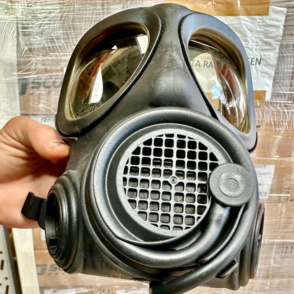 En delad bild där vänstra delen visar en hand som håller i en svart gasmask framför en bakgrund av kartonger. På den högra delen står Max Rupla, beredskapschef Region Västmanland, i en orange jacka i en lagerlokal.