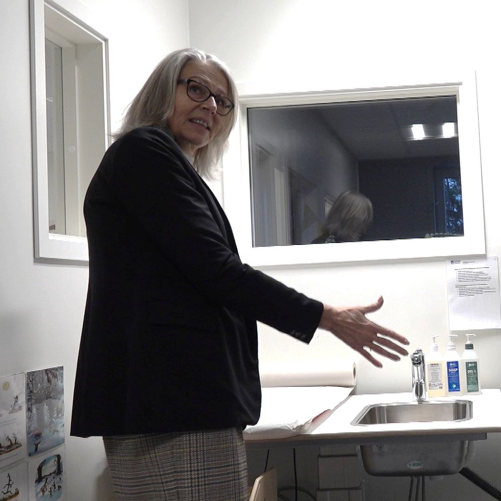 En kvinna står vid ett skrivbord i ett kontorsrum och gestikulerar med sin hand. I rummet finns en handfat och hyllor fyllda med olika förnödenheter, samt några dokument som hänger på väggen. Rummet verkar ljust och välordnat.