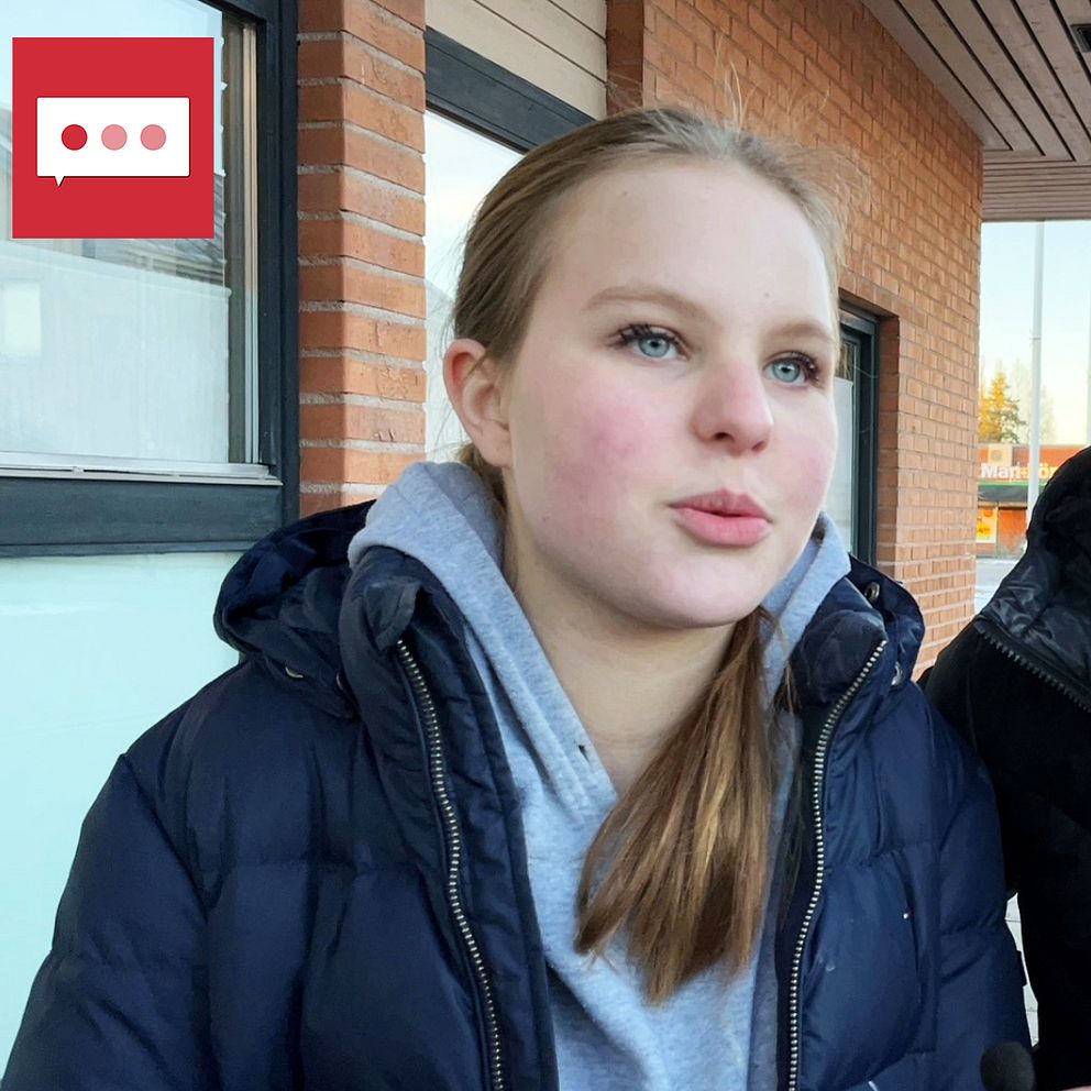 Två personer står tillsammans utomhus, iförda vinterjackor. Bakgrunden visar en byggnad och en klar blå himmel. De verkar vara involverade i ett samtal eller en aktivitet.